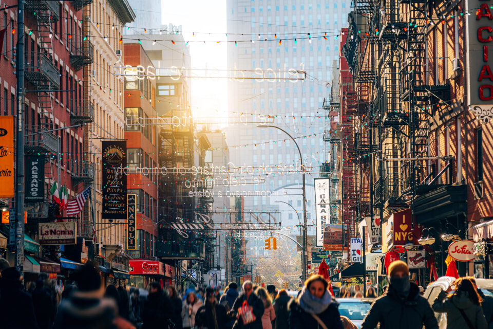 A lively street in Manhattan surrounded by historic buildings, colorful storefronts, and numerous pedestrians enjoying an urban walk. The image captures the vibrant urban life of New York City.