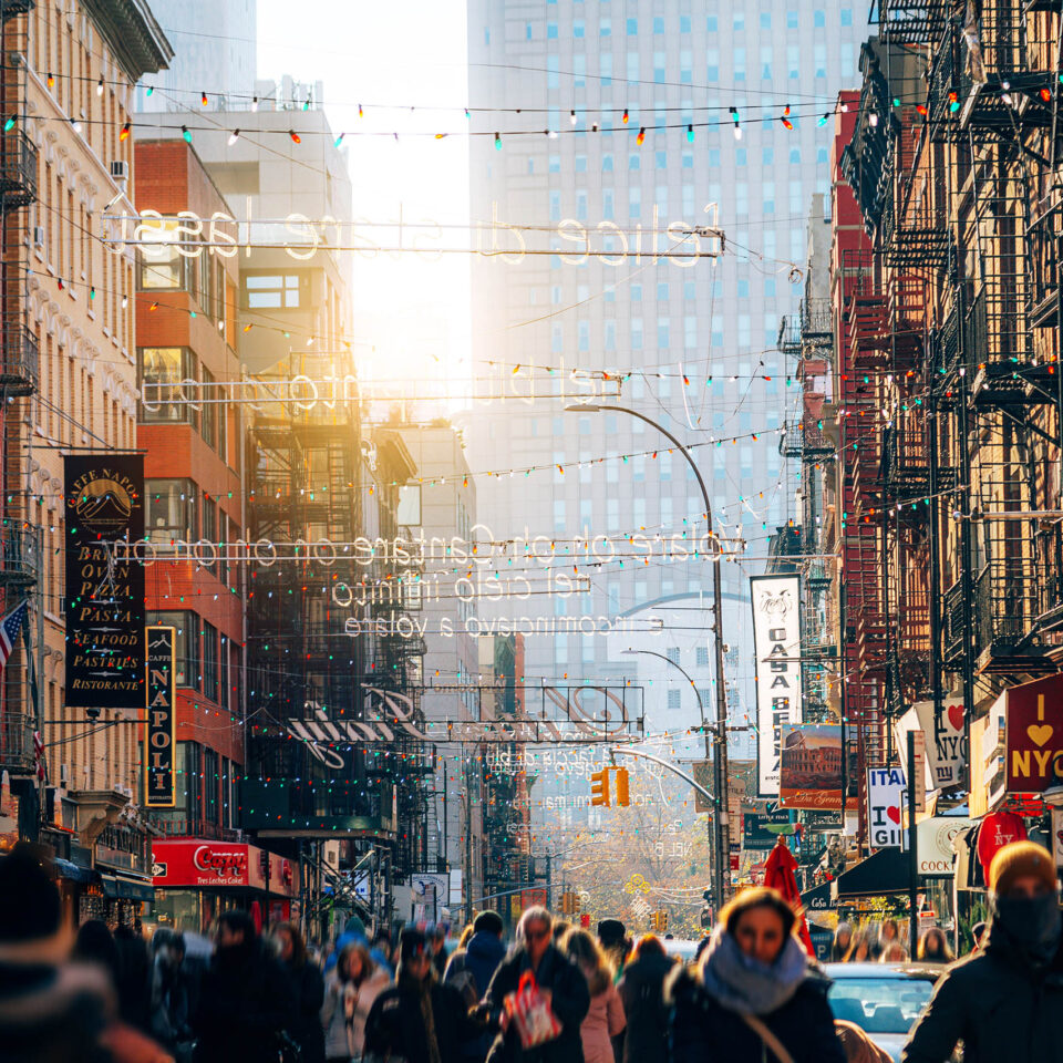 A lively street in Manhattan surrounded by historic buildings, colorful storefronts, and numerous pedestrians enjoying an urban walk. The image captures the vibrant urban life of New York City.