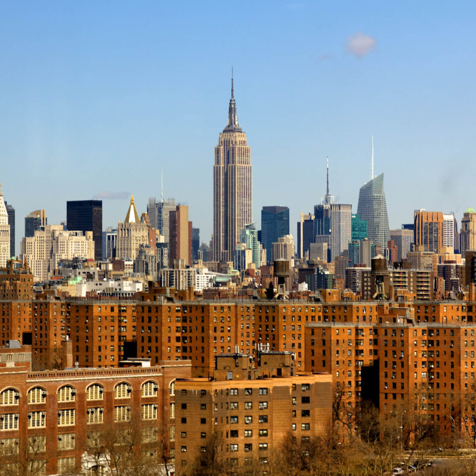 The New York City midtown Manhattan skyline with the landmark Empire State Building in the center and contrasting drab brown apartment blocks in the foreground. Shot from across the East River in early spring.