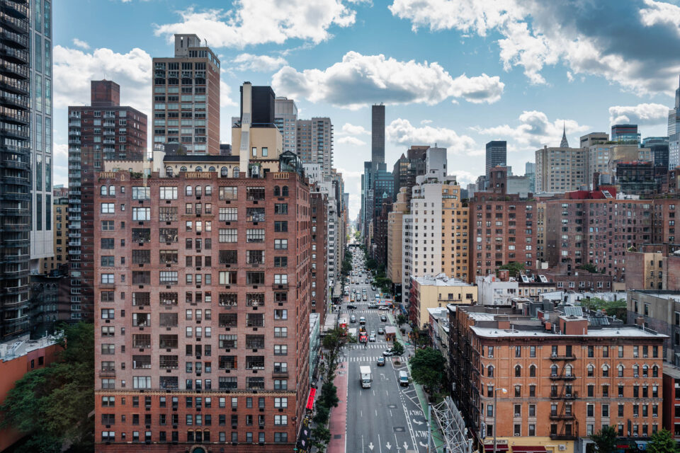Urban cityscape view along Second Avenue, showing dense residential and commercial buildings, traffic, and classic Midtown East architecture under a partly cloudy sky. The image conveys everyday city life, real estate density, and metropolitan scale in New York City, Upper East Side, Manhattan, New York, United States, North America.