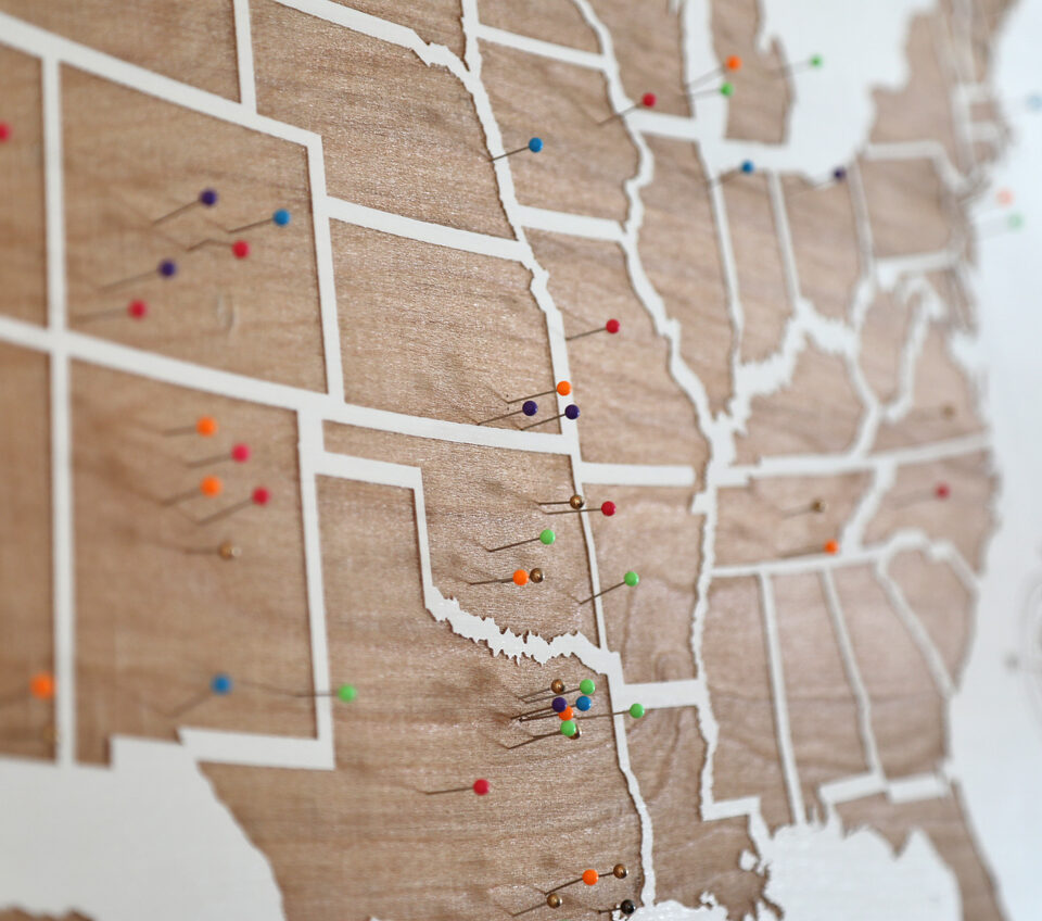 Wooden US States map on the wall with multicolored pins on it.